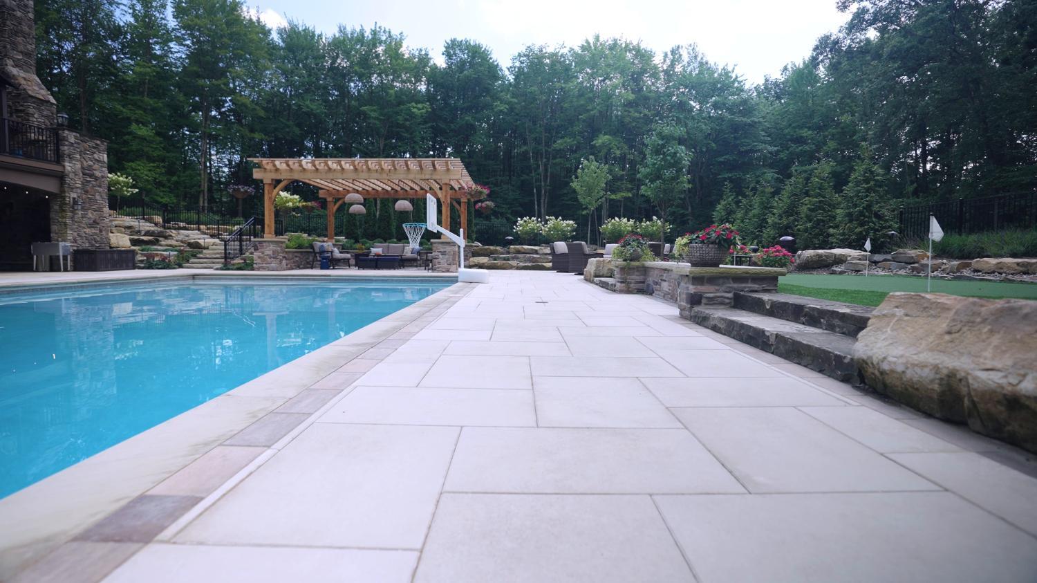 Long pool with a stone deck and pergola in a tree-lined backyard