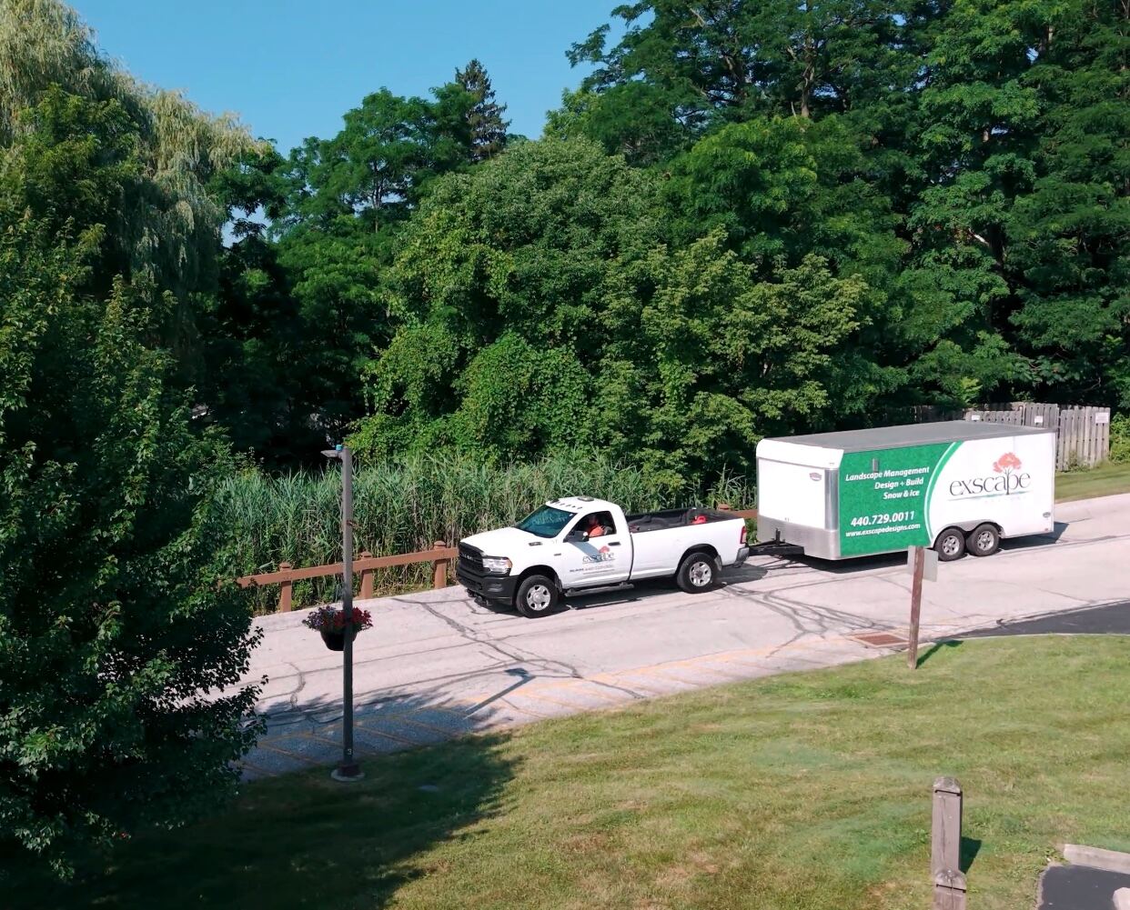 Exscape Designs landscaping company truck towing a branded trailer on a suburban road surrounded by trees and greenery