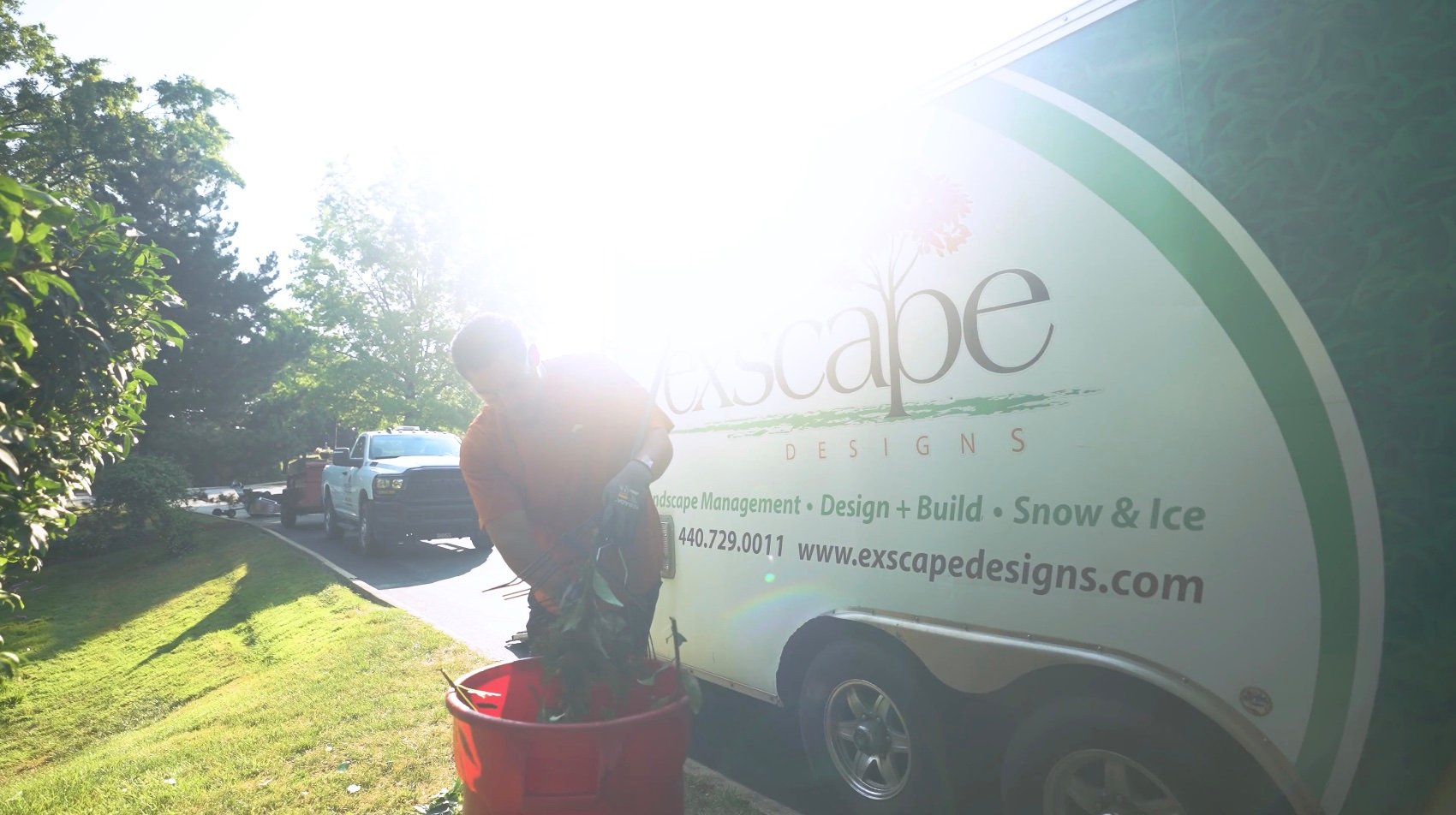 Exscape Designs landscaping crew member loading debris into a bin beside company trailer and truck during a maintenance project in Northeast Ohio.