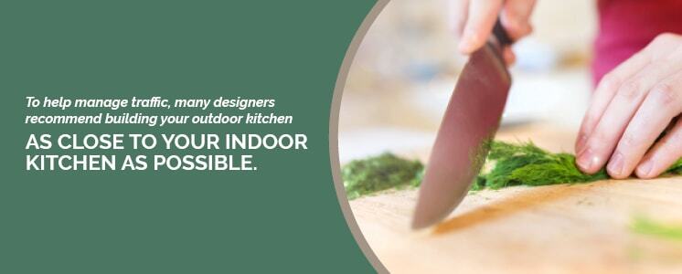 A very close up shot of a cutting board with a hands holding a large knife cutting herbs cropped in a circle with text to the left reading "To help manage traffic, many designers recommend building your outdoor kitchen as close to your indoor kitchen as possible."