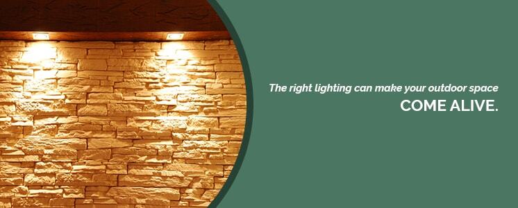 Two very warm toned lights set to shine down on a light tan wall making it appear very bright in a dim setting cropped in a circle with text to the left reading "The right lighting can make your outdoor space come alive."