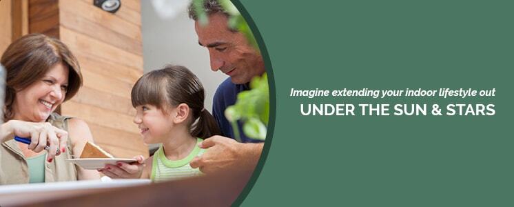 From a low angle partially obscured by a wooden table, a family of three with the mother holding and serving a perfect slice of cake to a roughly 8 year old girl with the father behind the young girl gently embracing her shoulders, all cropped into a circle with text to the right reading "Imagine extending your indoor lifestyle out under the sun & stars"