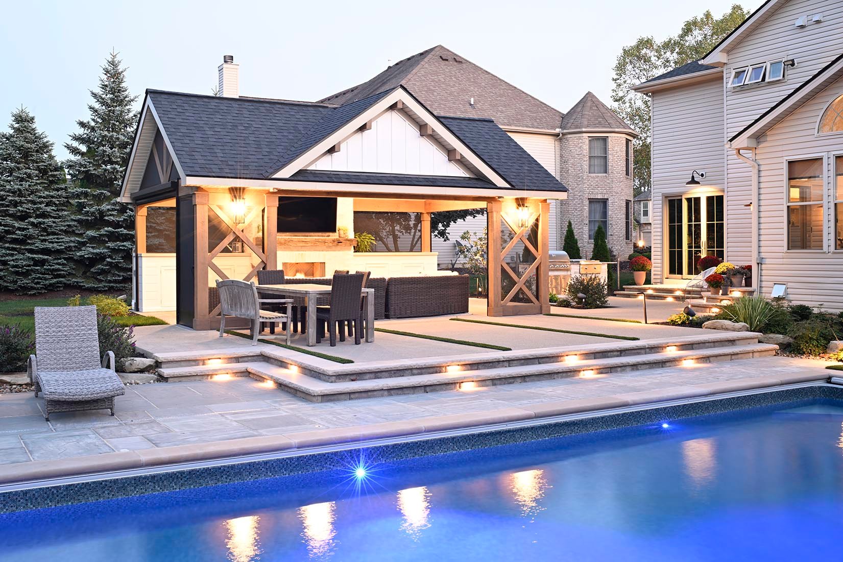 Pool house with outdoor kitchen beside a lit inground pool